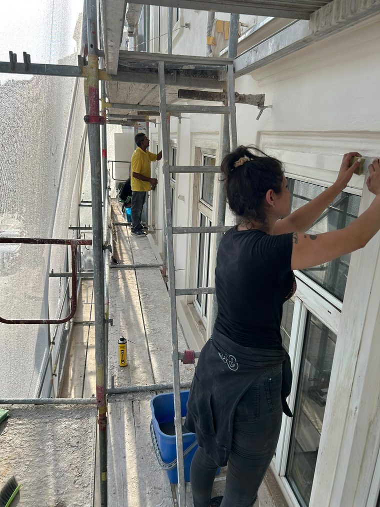 Zwei Reinigungskräfte reinigen Fensterrahmen einer frisch sanierten Altbaufassade von einem Baugerüst aus