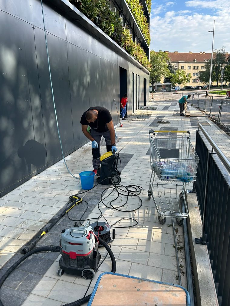 Reinigungskraft rüstet einen Hochdruckreiniger vor einer dunklen Metallfassade, daneben ein Nasssauger