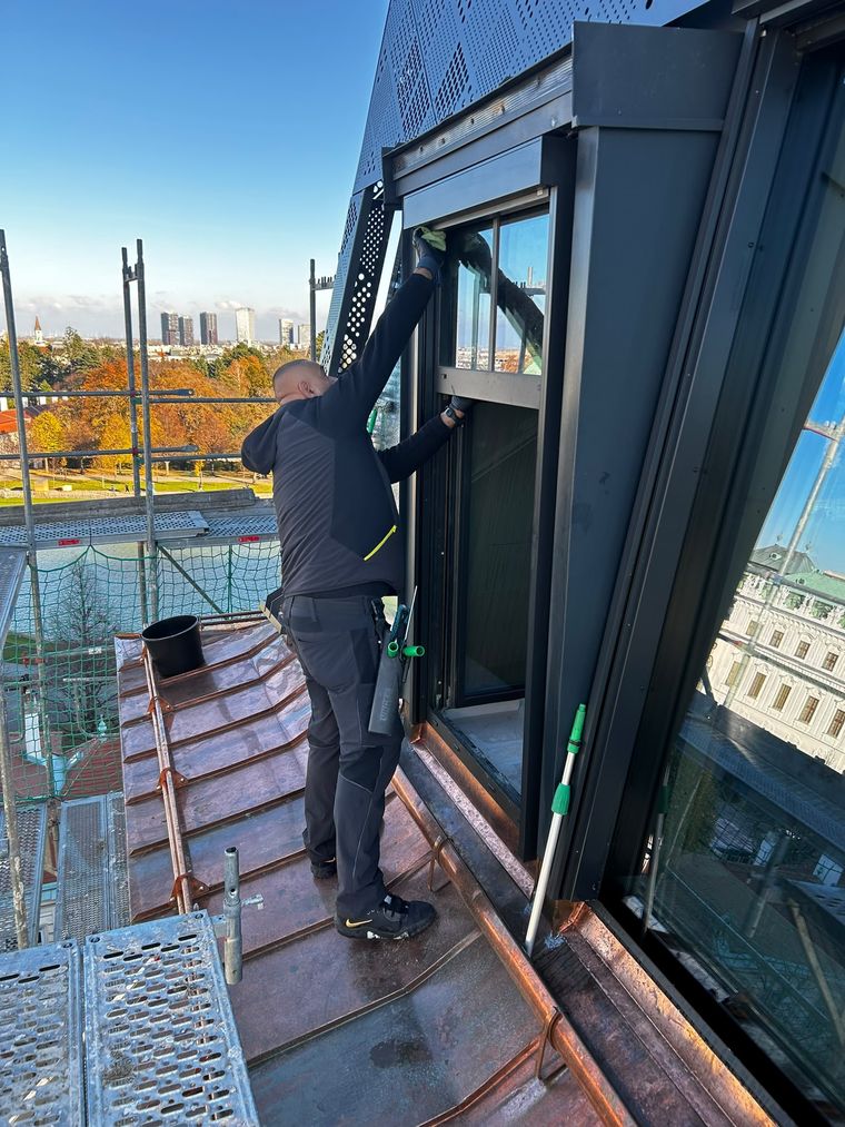 Reinigungskraft reinigt ein Dachflächenfenster auf einem Wiener Dach, gesichert über das Gerüst