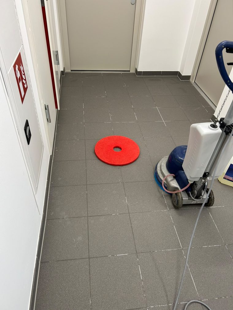 Gereinigter Gang mit grauem Fliesenboden, abgestellte Einscheibenmaschine und gebrauchtes rotes Reinigungspad