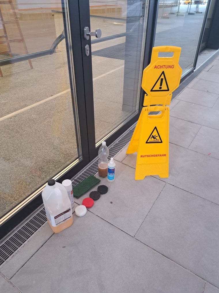 Reinigungspads, Reinigungsmittel und eine Flasche mit Testwasser neben einem Warnschild auf Steinplatten