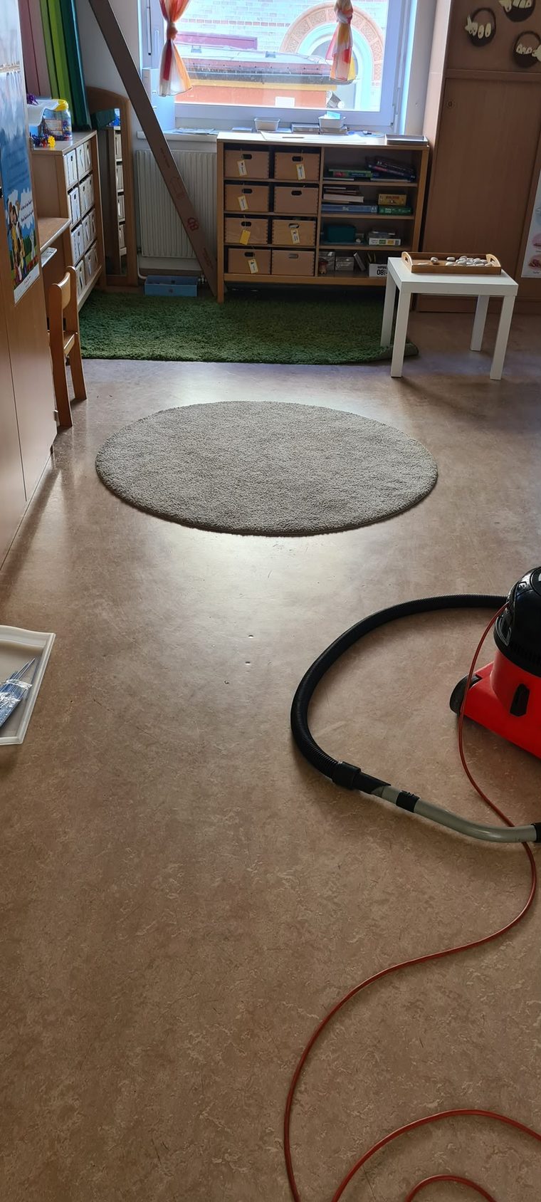 Gruppenraum mit rundem Teppich, Regalen und einem Staubsauger mit Schlauch auf dem Hartboden